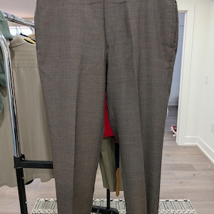 May include: A pair of brown trousers hanging on a wooden hanger. The trousers are made of a textured fabric and feature a button closure and pockets. The trousers are displayed against a neutral background.