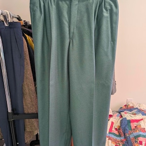 Puede incluir: Pantalones plisados de color verde azulado con una etiqueta de talla 32. Los pantalones están colgados de una percha. Se ve otra ropa en el fondo. La tela parece ser de peso medio.