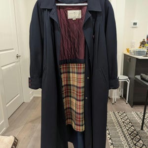 May include: A navy blue trench coat hanging on a white hanger. The coat has a burgundy quilted lining and a plaid pattern on the lower interior. The coat has a classic collar, button closures, and long sleeves.