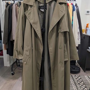 Könnte beinhalten: Ein langer, olivgrüner Trenchcoat mit breitem Kragen und Knopfdetails. Der Mantel hängt an einem schwarzen Kleiderbügel und zeigt seine volle Länge und sein Design. Der Mantel ist aus strapazierfähigem Stoff gefertigt, der für verschiedene Wetterbedingungen geeignet ist.