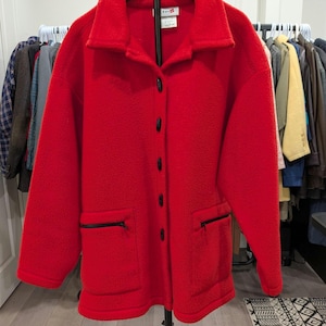 May include: A bright red fleece jacket featuring a collared neckline, a full-length button-down front closure with black buttons, and two front pockets with black zippered openings. The jacket has long sleeves and a straight hemline.