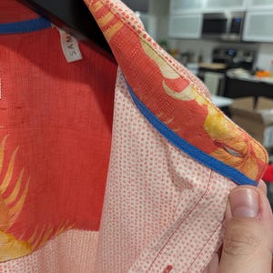 May include: Close-up of a coral-colored button-down shirt with a floral pattern on the collar and cuffs. The collar is trimmed with a blue ribbon. The shirt has a dotted pattern on the front.
