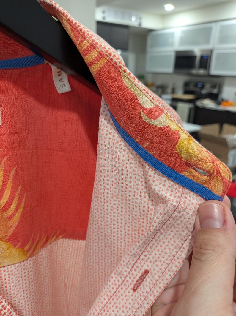 May include: Close-up of a coral-colored button-down shirt with a floral pattern on the collar and cuffs. The collar is trimmed with a blue ribbon. The shirt has a dotted pattern on the front.