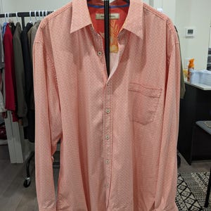 May include: Coral-colored button-down shirt with a small white dotted pattern. The shirt has long sleeves, a chest pocket, and a patterned interior collar. It is displayed on a black hanger.