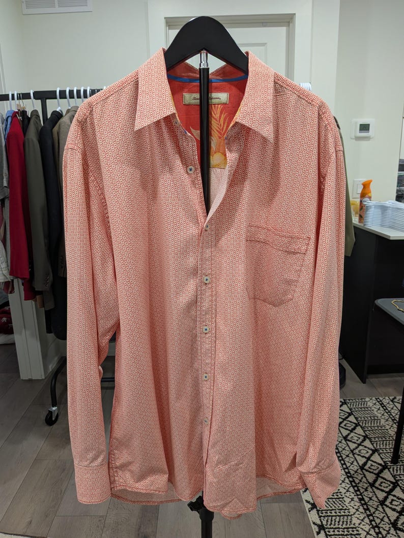 May include: Coral-colored button-down shirt with a small white dotted pattern. The shirt has long sleeves, a chest pocket, and a patterned interior collar. It is displayed on a black hanger.