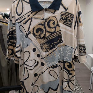 May include: A short-sleeved polo shirt with a navy blue collar and a button-down front. The shirt features a beige, black, and light blue abstract pattern. The shirt is hanging on a white hanger.
