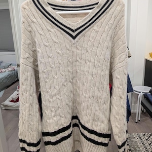 May include: Cream-colored cable knit sweater with a V-neck and black stripes. The sweater has a classic design with black stripes at the neckline and hem. The sweater is hanging on a white hanger.