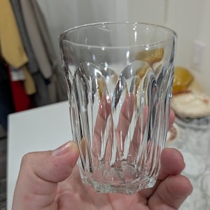 May include: A clear, vintage-style drinking glass with a faceted design. The glass has a thick base and a slightly flared rim. The sides feature vertical, curved grooves that catch the light. This glassware is suitable for water, juice, or cocktails.
