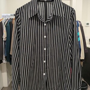 May include: A long-sleeved button-down shirt featuring a vertical pinstripe pattern in black and white. The shirt has a classic pointed collar, a full-length front button placket with white buttons, and buttoned cuffs on the long sleeves.