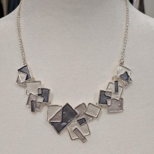 May include: Silver necklace with a geometric design. The necklace features interconnected square and rectangular shapes in grey and black. The chain is silver, and the pendant is displayed on a white mannequin. The necklace is a modern piece of jewellery.