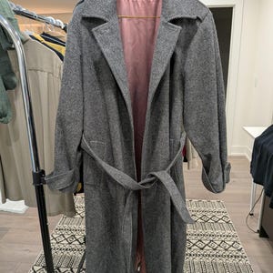 Peut inclure: Un long manteau gris avec une ceinture assortie, suspendu à un portant. Le manteau a un design classique avec un col cranté et une doublure rose. Le tissu présente un motif subtil et le manteau semble être fait d'une matière de poids moyen.