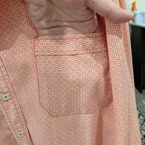 May include: A button-down shirt in a coral color with a small white dot pattern. The shirt has a chest pocket and blue buttons. The fabric appears to be lightweight.