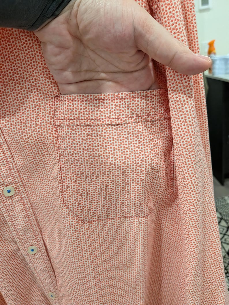 May include: A button-down shirt in a coral color with a small white dot pattern. The shirt has a chest pocket and blue buttons. The fabric appears to be lightweight.