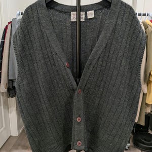 Può includere: Un gilet senza maniche grigio scuro con motivo a maglia a coste verticali, scollo a V e chiusura frontale con quattro bottoni bordeaux. Il capo presenta un'etichetta all'interno del colletto con la scritta "THE TAILORS".
