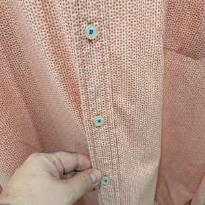 May include: Close-up of a button-down shirt in a peach and white dotted pattern. The shirt has a classic collar and a buttoned placket with four visible buttons. The buttons are off-white with a blue accent.