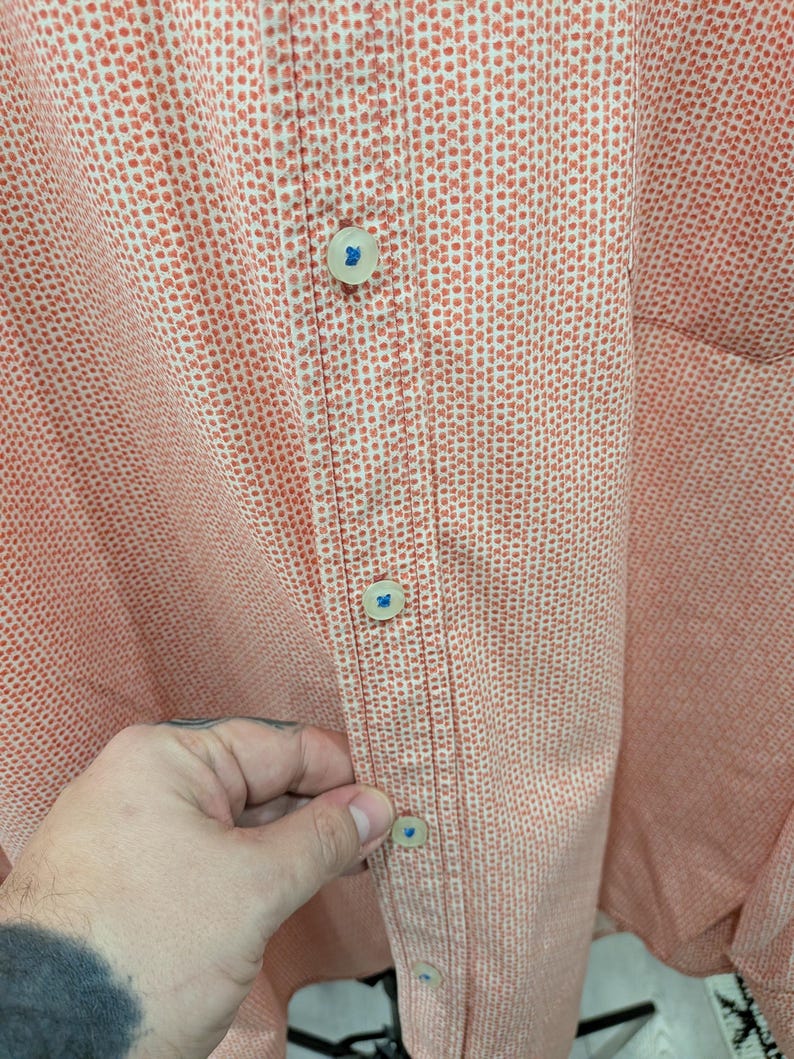 May include: Close-up of a button-down shirt in a peach and white dotted pattern. The shirt has a classic collar and a buttoned placket with four visible buttons. The buttons are off-white with a blue accent.