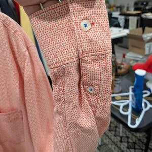 May include: Close-up of a coral-colored long-sleeve shirt sleeve with a white dotted pattern. The cuff features two blue buttons. The shirt is made of a textured fabric.