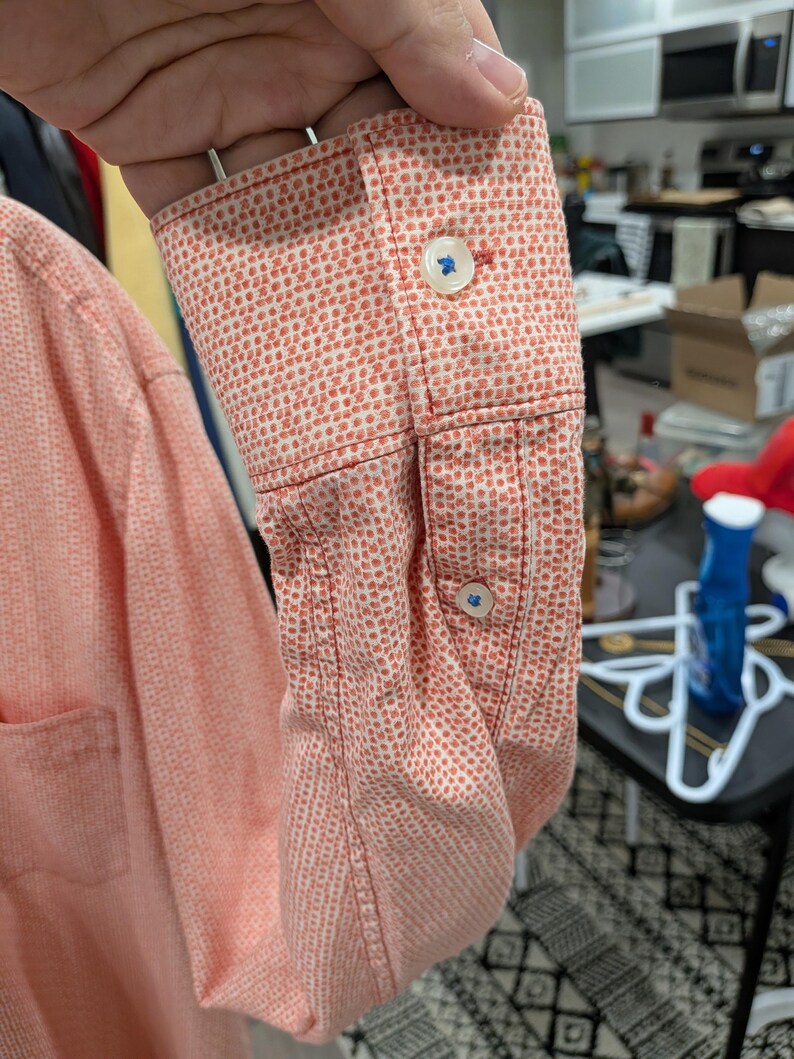 May include: Close-up of a coral-colored long-sleeve shirt sleeve with a white dotted pattern. The cuff features two blue buttons. The shirt is made of a textured fabric.