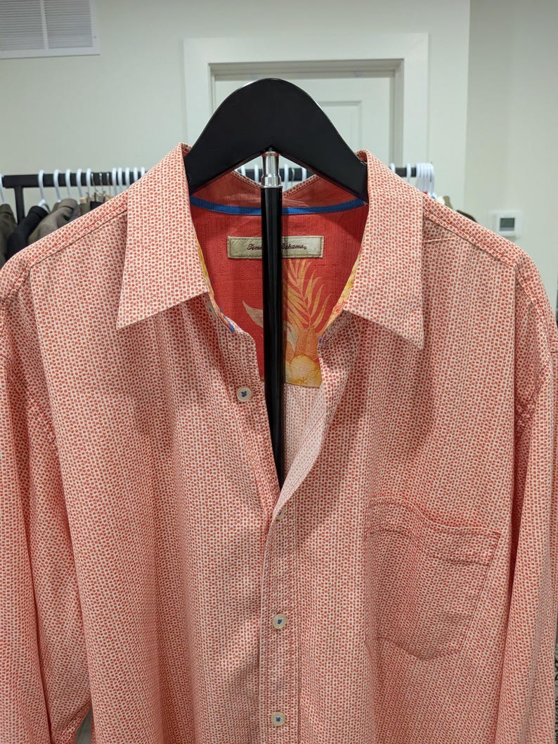 May include: Coral-colored button-down shirt with a small dot pattern. The shirt has a chest pocket and a patterned interior lining. The shirt is hanging on a black hanger.