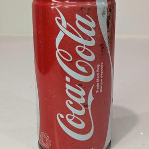 May include: A red and white aluminium Coca-Cola beverage can with a 280 ml capacity. The can features the white Coca-Cola logo, the text Trade Mark Reg. Marque déposée, and a small white circular icon at the bottom. The surface shows signs of wear.