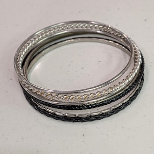 Puede incluir: Un conjunto de pulseras plateadas y negras. Las pulseras presentan diversas texturas, lisas, trenzadas y con forma de cuerda. Las pulseras están apiladas, creando un efecto de capas. La paleta de colores es principalmente plateada y negra.