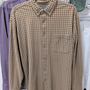 May include: A brown and cream plaid long-sleeved shirt with a button-down collar and a chest pocket. The shirt is displayed on a white hanger. Other shirts in purple and white are visible in the background.