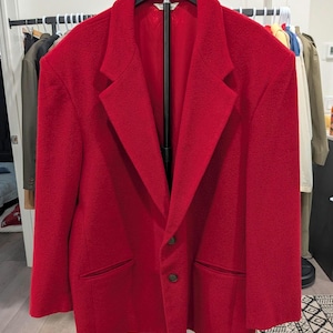 May include: A vibrant red blazer with a classic two-button closure and notched lapels. The jacket features two front pockets and is displayed on a black hanger. The blazer is made of a textured fabric.