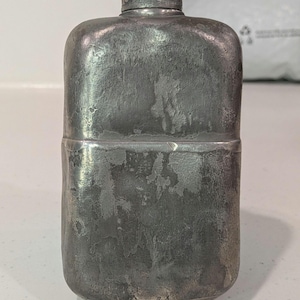 May include: A rectangular metal flask with a weathered, dark grey finish and a textured, uneven surface. The flask features a central horizontal seam dividing the body, rounded corners, and a small, cylindrical screw-top cap at the centre top.