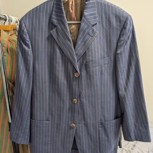 May include: Blue blazer with vertical tan stripes, three gold-colored buttons, and two front pockets. The jacket is hanging on a hanger. The blazer has a classic design.