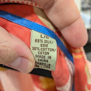 May include: Close-up of a tag from a silk and cotton shirt. The tag is green with black text that reads "L/G 65% SILK/ SOIE 35% COTTON/ COTON MADE IN CHINA FABRIQUE EN CHINE". The shirt has a colorful floral pattern.