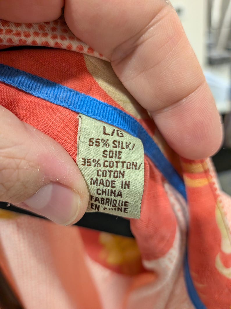 May include: Close-up of a tag from a silk and cotton shirt. The tag is green with black text that reads "L/G 65% SILK/ SOIE 35% COTTON/ COTON MADE IN CHINA FABRIQUE EN CHINE". The shirt has a colorful floral pattern.