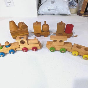 May include: A collection of wooden toy train carriages featuring natural wood finishes and colourful accents. The set includes various engine and carriage shapes with small wooden wheels in blue, red, green, and yellow, designed for modular assembly.