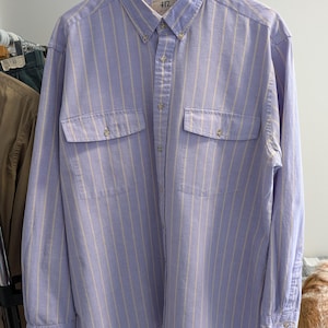 Puede incluir: Una camisa abotonada con rayas verticales en lila claro y crema. La camisa de manga larga tiene dos bolsillos en el pecho y un cuello abotonado. La etiqueta dice "417". La camisa está colgada en una percha.