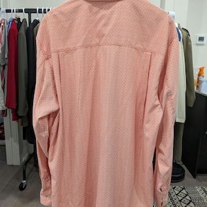 May include: A long-sleeved button-down shirt in a salmon color, patterned with small, repeating red dots. The shirt is hanging on a black hanger, with other clothing visible in the background.