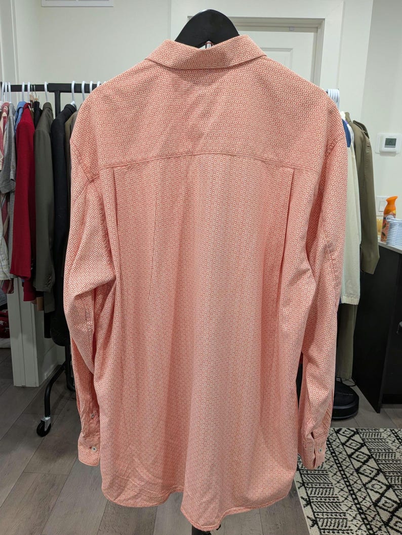 May include: A long-sleeved button-down shirt in a salmon color, patterned with small, repeating red dots. The shirt is hanging on a black hanger, with other clothing visible in the background.