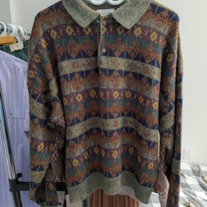 May include: A patterned sweater with a collared neckline and button closure. The sweater displays a geometric design in brown, blue, yellow, and green hues. The collar and cuffs are a solid olive green. The garment is suspended on a white hanger.