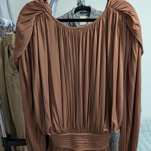 May include: A long-sleeved, brown dress with a gathered bodice and a cinched waist. The dress features a scoop neckline and long sleeves with gathered detailing at the shoulders. The dress is hanging on a black hanger.