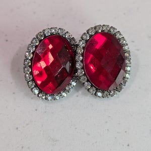 Puede incluir: Un par de pendientes de botón ovalados. Cada pendiente presenta una gema facetada de color rojo intenso rodeada por un halo de pequeños cristales transparentes. Los pendientes están engarzados en metal plateado.