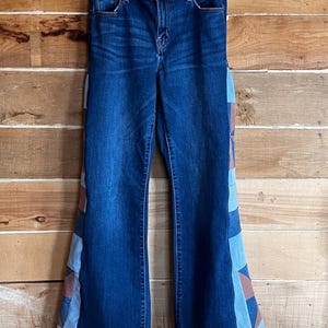 May include: Dark wash denim jeans with a high waist and flared legs. The sides of the jeans feature a patchwork design with geometric shapes in shades of blue and brown. The jeans are hanging on a metal hanger against a wooden background.