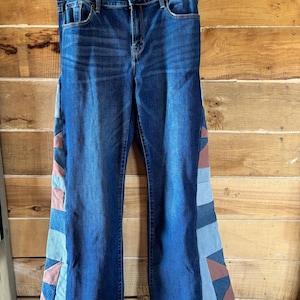 Könnte beinhalten: Dunkelblaue Jeans mit Patchwork-Details an den ausgestellten Beinen. Das Patchwork zeigt geometrische Formen in Blau-, Rosa- und Brauntönen. Die Jeans hängen an einem transparenten Kunststoffbügel.