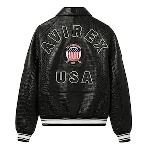 Puede incluir: Chaqueta de cuero negro con un patr&oacute;n texturizado similar al de un cocodrilo. Presenta la palabra "AVIREX" arqueada sobre los hombros, con "USA" debajo de un emblema circular. El emblema incluye las palabras "ALL AMERICANS" y "ESTABLISHED 1975". Pu&ntilde;os y dobladillo a rayas blancas y azul marino.