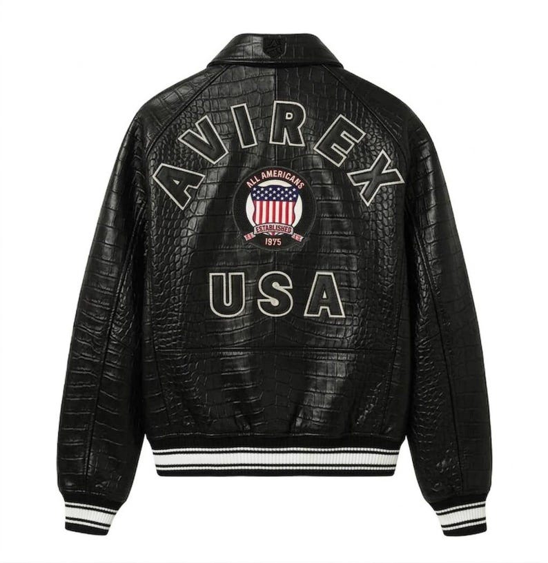 Puede incluir: Chaqueta de cuero negro con un patr&oacute;n texturizado similar al de un cocodrilo. Presenta la palabra "AVIREX" arqueada sobre los hombros, con "USA" debajo de un emblema circular. El emblema incluye las palabras "ALL AMERICANS" y "ESTABLISHED 1975". Pu&ntilde;os y dobladillo a rayas blancas y azul marino.