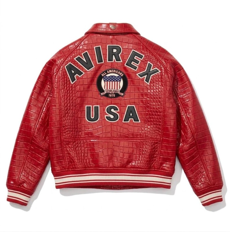May include: A red leather jacket with a crocodile skin effect. The back displays the text "AVIREX" and "USA" in white, with a circular patch featuring the American flag. The jacket has a white and black striped trim on the cuffs and hem.