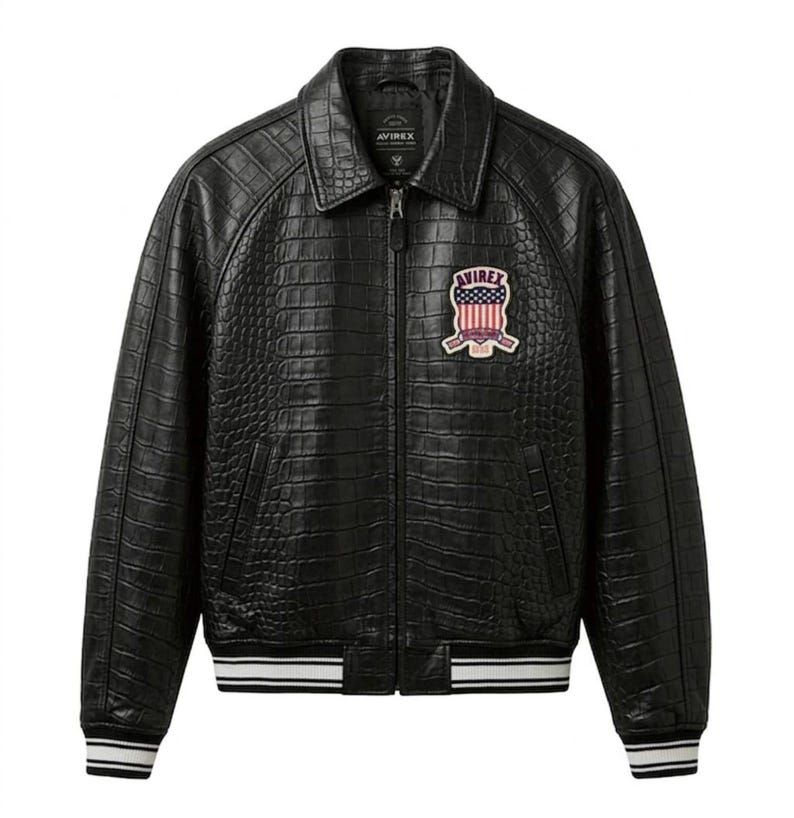 Puede incluir: Chaqueta de cuero negro con textura de cocodrilo. La chaqueta tiene cuello, cierre de cremallera y un parche en el pecho con el texto "AVIREX". Los pu&ntilde;os y el dobladillo tienen detalles a rayas blancas y negras.