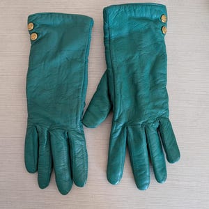 May include: A pair of emerald green leather gloves. Each glove has three gold-colored buttons at the wrist. The gloves are long, extending past the wrist, and have a textured appearance.
