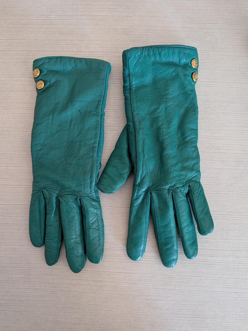 May include: A pair of emerald green leather gloves. Each glove has three gold-colored buttons at the wrist. The gloves are long, extending past the wrist, and have a textured appearance.