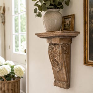 May include: A wall-mounted wooden shelf featuring a carved corbel support with leaf motifs. The shelf holds a light-colored ceramic vase containing green foliage and a small framed landscape painting, showcasing a weathered brown wood finish.