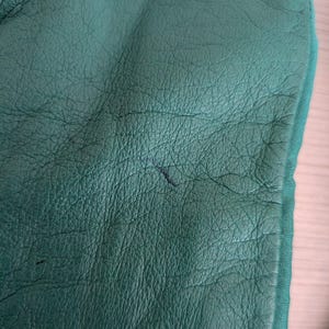 May include: Close-up of a textured, emerald green leather surface. The leather has a fine grain and visible wrinkles. A darker mark is visible on the surface. The edge of a green fabric is visible.