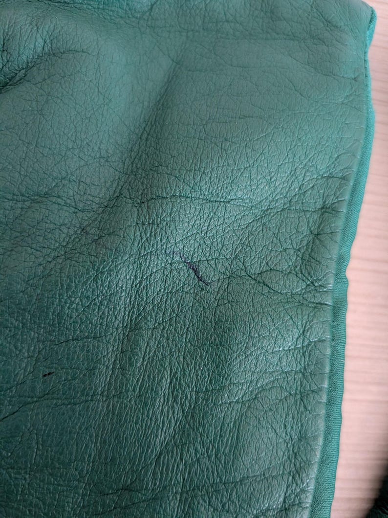May include: Close-up of a textured, emerald green leather surface. The leather has a fine grain and visible wrinkles. A darker mark is visible on the surface. The edge of a green fabric is visible.