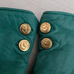 May include: Close-up of a pair of emerald green leather gloves. Each glove has three gold-colored buttons with a stylized arrow design. The gloves are made of a soft, textured leather.
